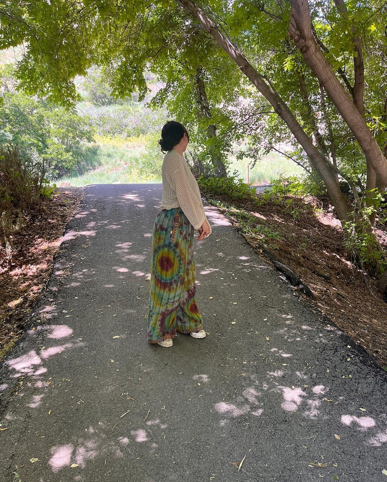 Puede incluir: Una persona con un top de crochet blanco y pantalones tie-dye se encuentra en un camino pavimentado en un &aacute;rea boscosa. El camino est&aacute; bordeado de &aacute;rboles y vegetaci&oacute;n verde.
