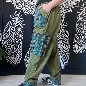 May include: Green, loose-fitting pants with a blue and gold patterned fabric panel on the left leg. The pants have a drawstring waist and a wide leg. The pants are being worn with black and white sneakers.