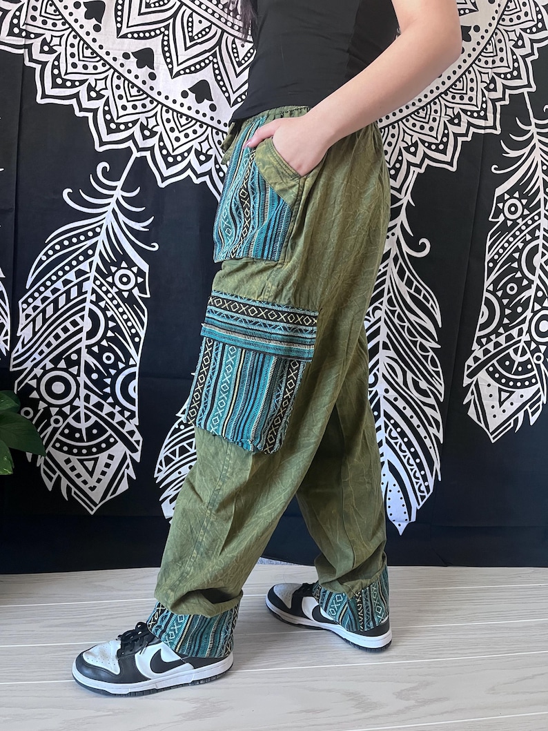 May include: Green, loose-fitting pants with a blue and gold patterned fabric panel on the left leg. The pants have a drawstring waist and a wide leg. The pants are being worn with black and white sneakers.