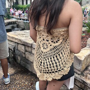May include: A beige crochet top with a scalloped hem. The top is sleeveless and has a loose, flowy fit.