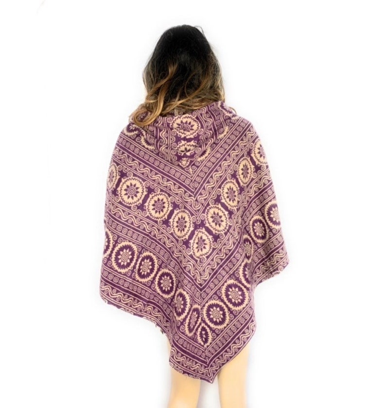 K&ouml;nnte beinhalten: Lilafarbener Kapuzenponcho mit einem cremefarbenen und lilafarbenen geometrischen Muster. Das Muster zeigt ein sich wiederholendes Design aus Kreisen und Spiralen.