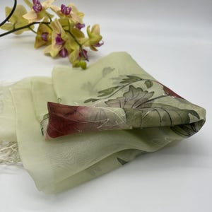 May include: A light green sheer scarf with a floral print in shades of brown, red, and white. The scarf has a fringed edge.