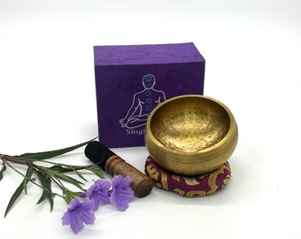 Puede incluir: Un cuenco tibetano de latón con un percutor de madera y un cojín morado. Una caja morada con una ilustración de una figura humana y la palabra "Singi" está detrás del cuenco. Flores moradas y hojas verdes en primer plano.