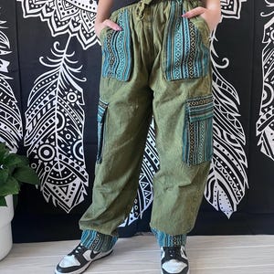 May include: Green cargo pants with blue and black patterned fabric panels. The pants have a drawstring waist and multiple pockets.