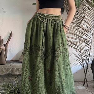 Pode incluir: Uma saia longa verde-oliva com bordados florais e de rolagem. A saia tem cintura com cordão e bainha com babados. Uma corrente dourada é usada na cintura. A pessoa está usando um top preto.