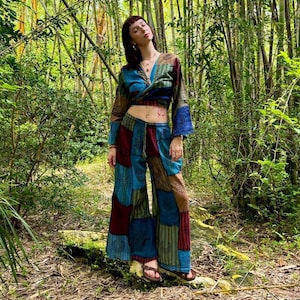 Puede incluir: Una mujer con un pantalón de vestir de color azul, rojo y verde con un top de manga larga y un escote de estilo envolvente. Está de pie en un bosque con árboles de bambú.