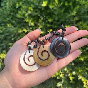 May include: Three wooden pendants with a spiral design. The pendants are on a black cord with brown wooden beads. The pendants are white, brown, and black.