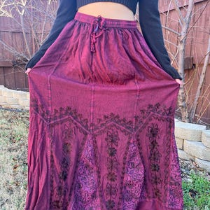 May include: A long, flowing burgundy skirt with an embroidered floral design. The skirt has a drawstring waist and a full, gathered silhouette. The embroidery is dark brown, creating a contrast against the burgundy fabric. The skirt is made of a soft, flowing material.