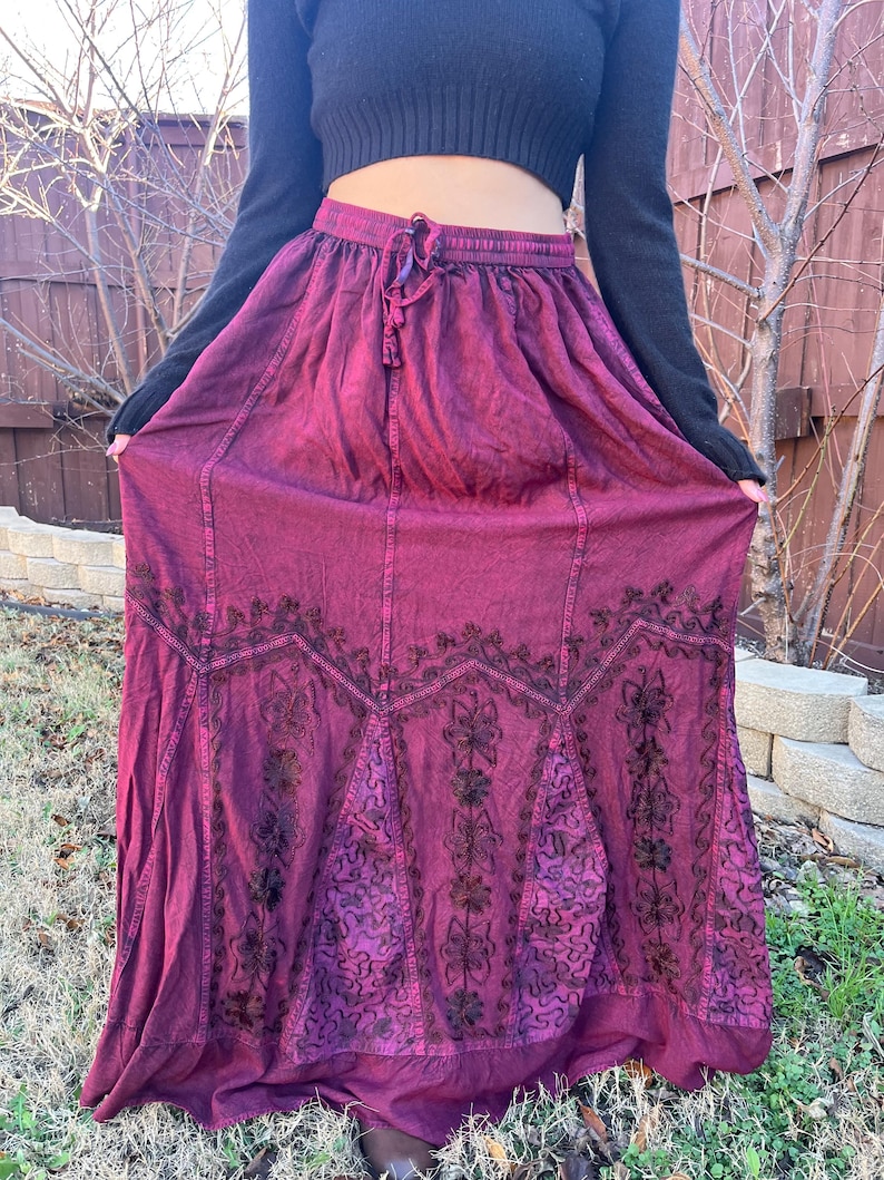 May include: A long, flowing burgundy skirt with an embroidered floral design. The skirt has a drawstring waist and a full, gathered silhouette. The embroidery is dark brown, creating a contrast against the burgundy fabric. The skirt is made of a soft, flowing material.