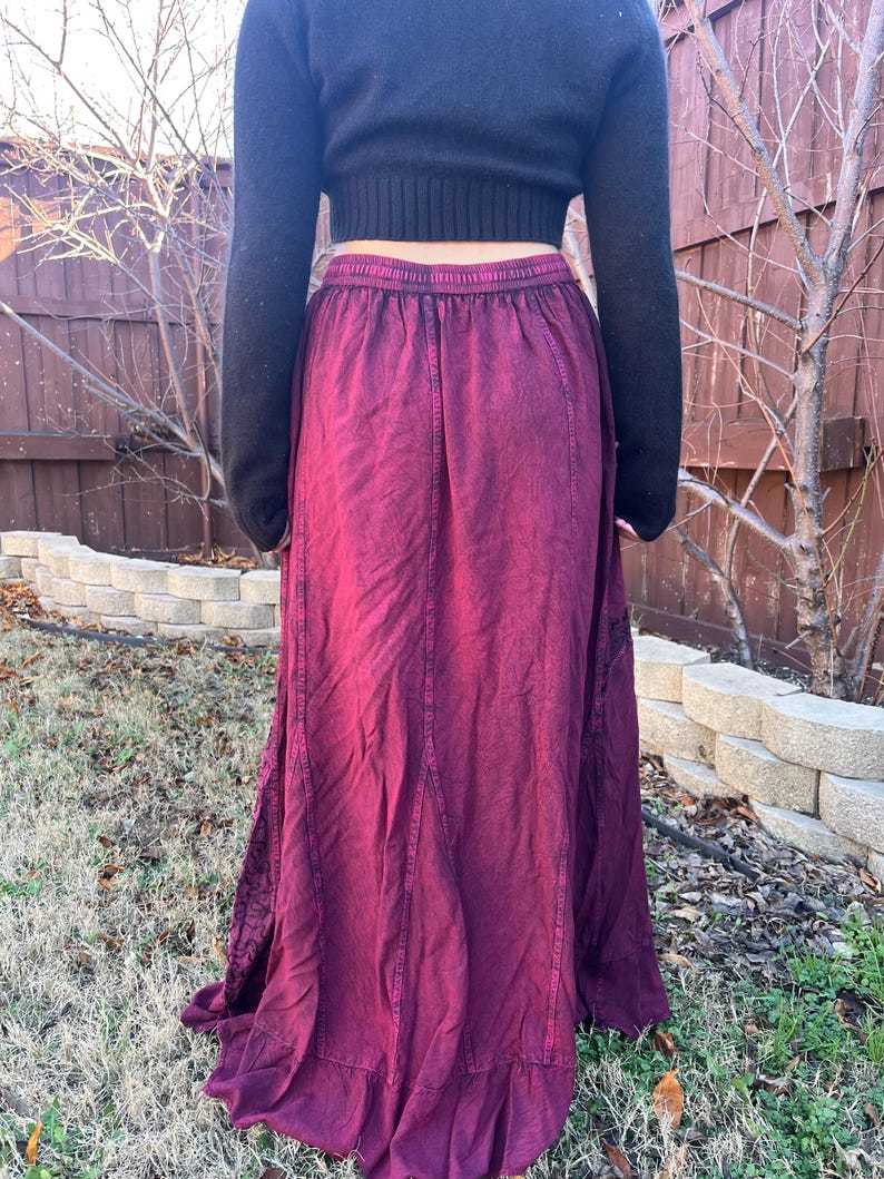 May include: A long, flowing burgundy skirt with a ruffled hem. The skirt has a high waist and a textured, slightly faded appearance. The person is wearing a black sweater.