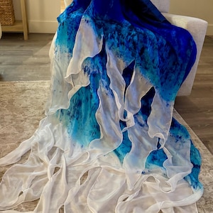 Puede incluir: Una bufanda fluida en tonos azul oscuro, turquesa y blanco. La bufanda está drapeada sobre una silla, mostrando su diseño en capas y con volantes. La tela parece ligera y aireada, con un efecto de acuarela.