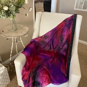 May include: A purple and red silk scarf with black accents draped over a white armchair. The scarf has a tie-dye pattern.