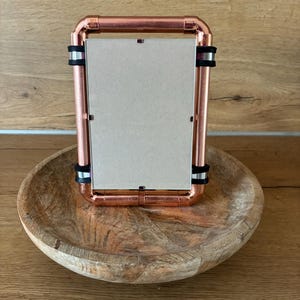 Pode incluir: Uma moldura retangular de metal cor de cobre. A moldura é mantida junta por bandas de borracha pretas. Está sobre uma tigela de madeira com um acabamento natural.