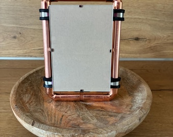 May include: A copper-colored metal picture frame with a rectangular shape. The frame is held together with black rubber bands. The frame is sitting on a wooden bowl with a natural finish.