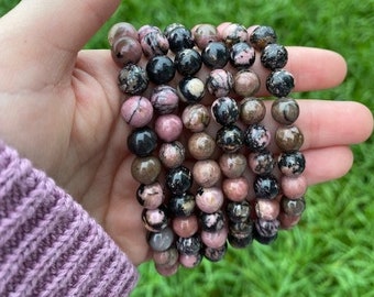 May include: A stack of bracelets made with pink and black patterned beads. The bracelets are made with round beads and are stacked on top of each other.