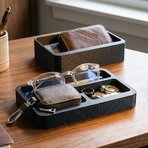 Puede incluir: Un escritorio de madera con una bandeja organizadora negra que contiene gafas, un llavero y monedas. Una cartera de cuero se encuentra en una bandeja similar. Una lámpara de escritorio de latón y una planta suculenta también están en el escritorio.