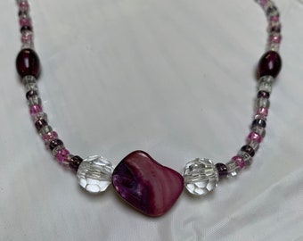 Puede incluir: Un collar de cuentas de concha púrpura y rosa con cuentas de vidrio transparentes y pequeñas cuentas púrpuras. El collar está sobre un fondo blanco.