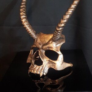 Könnte beinhalten: Eine metallisch bronzefarbene Totenkopfmaske mit zwei langen, geschwungenen und gedrehten Hörnern, die sich von der Stirn nach oben erstrecken. Die Maske hat tiefe Augenhöhlen, eine Nasenhöhle und einen detaillierten Kiefer mit sichtbaren Zähnen.