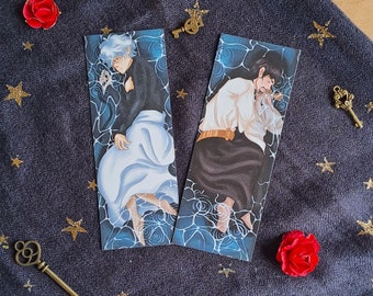 May include: Two blue and white illustrated bookmarks featuring a sleeping person with a white shirt and black trousers and a sleeping person with a white shirt and black trousers. The bookmarks are on a dark blue fabric background.