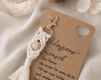 Op de afbeelding: Een handgemaakte macramé sleutelhanger met een houten hartbedel en een kwastje. De sleutelhanger is bevestigd aan een kaart met de tekst "Herzensmensch" en een hartvormige uitsparing. Een kandelaar en een gedroogde plant staan ook in de afbeelding.