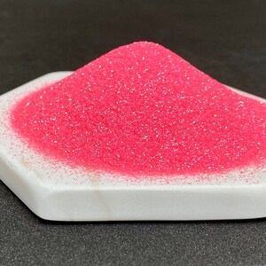 May include: A pile of pink glitter with a fine, iridescent shimmer. The glitter is displayed on a white hexagonal platform.