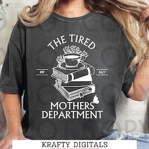 May include: A dark gray t-shirt with a white graphic design. The graphic features a stack of books with a cup of coffee and flowers on top. The text "The Tired Mothers Department" is below the books. The text "EST" and "24/7" are on either side of the books. The text "Krafty Digitals" is at the bottom of the image. The text "Instant Download PNG-300DPI Sublimation" is below the text "Krafty Digitals".