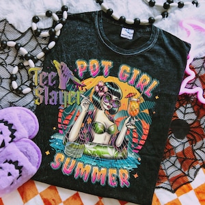 May include: Dark grey t-shirt with a colourful graphic of a woman in sunglasses holding a cocktail and a joint, with the words "Pot Girl Summer" and "Tee Slayer". The design features tropical elements.