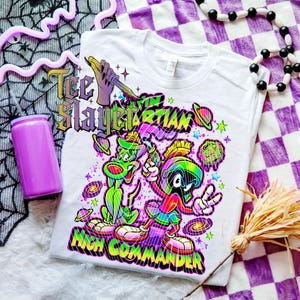 Op de afbeelding: Wit T-shirt met een kleurrijke afbeelding van twee cartoonaliens en de tekst "Tee Slayer Martian High Commander". Het ontwerp bevat planeten en sterren. Een paarse beker en decoratieve items zijn ook zichtbaar.