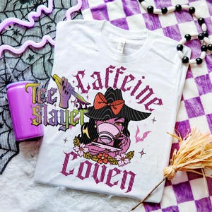 May include: White t-shirt with a graphic of a witch drinking coffee, the words "Caffeine Coven" and "Tee Slayer". The witch wears a black hat with a red bow. A purple tumbler with a straw is on the left.