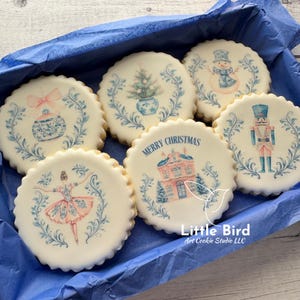 May include: A selection of festive sugar cookies, each with a unique Christmas design. The cookies are round and decorated with images of a snowman, Christmas tree, ballerina, and a house with the words "Merry Christmas". The designs use blue and pink colours.