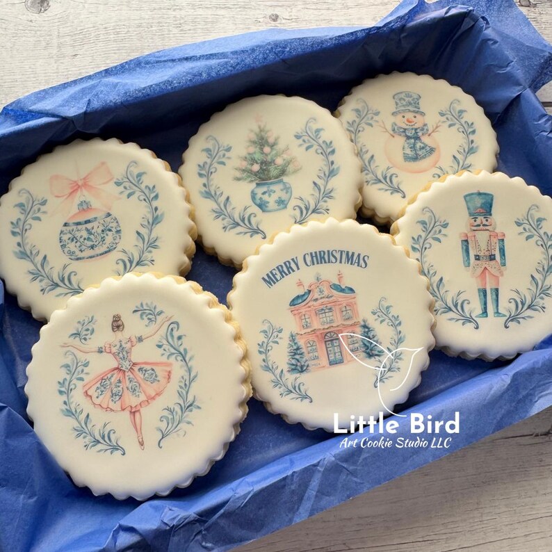 May include: A selection of festive sugar cookies, each with a unique Christmas design. The cookies are round and decorated with images of a snowman, Christmas tree, ballerina, and a house with the words "Merry Christmas". The designs use blue and pink colours.