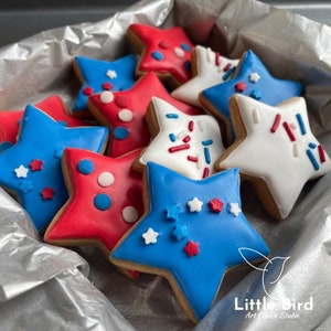 May include: A collection of star-shaped sugar cookies decorated in red, white, and blue icing. The cookies are arranged in a silver gift box. The cookies are decorated with sprinkles, dots, and stars.