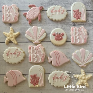 May include: A collection of decorated sugar cookies in a sea life theme. The cookies are in the shapes of a turtle, starfish, seashells, fish, and a crab. Some cookies have the words "BABY" and "baby price" in white icing. The cookies are pink, white, and beige.