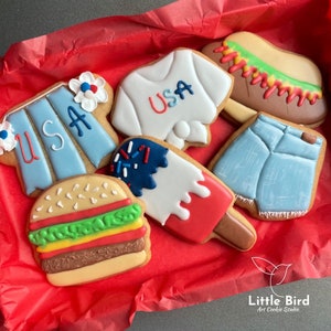 May include: A set of six decorated sugar cookies in a red gift box. The cookies are shaped like a hamburger, hot dog, popsicle, t-shirt, and two pieces of clothing. The cookies are decorated with red, white and blue icing and sprinkles. The t-shirt has the word "USA" written on it.