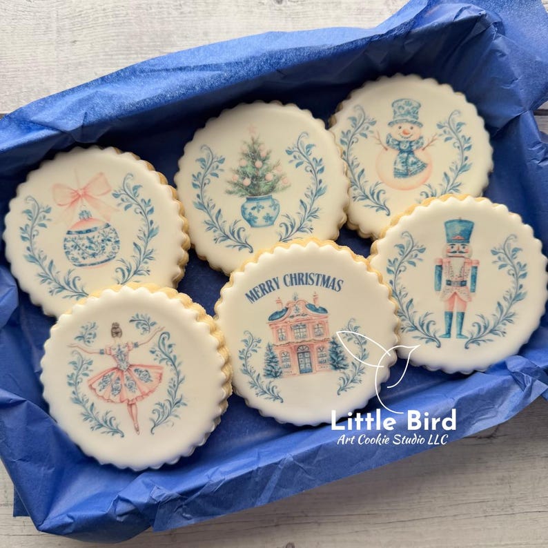 May include: Six round iced biscuits with scalloped edges, presented in a blue box. Each biscuit is decorated with a different Christmas design in blue and pink, such as a snowman, a nutcracker, and a ballerina. One biscuit has the text "MERRY CHRISTMAS".