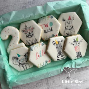 Peut inclure: Une boîte de biscuits en sucre décorés sur le thème des animaux de la jungle. Les biscuits sont hexagonaux et représentent un lion, un zèbre, une girafe, un éléphant, un rhinocéros et un numéro deux. La boîte est doublée de papier de soie bleu clair et porte une étiquette blanche avec le texte "Little Bird Art Cookie Studio LLC".