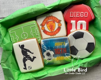 Könnte beinhalten: Eine Auswahl an dekorierten Zuckerkeksen mit Fußball-Motiven. Designs umfassen ein Fußballfeld, einen Fußball, ein Trikot mit "Diego" und der Nummer 10, ein Manchester United Logo und einen Fußballspieler. Die Kekse liegen in einer grün ausgekleideten Box.
