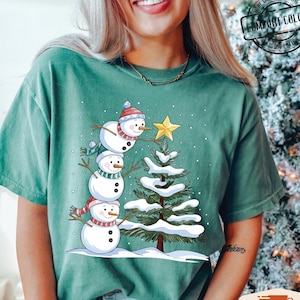 以下が含まれることがあります： 雪だるまの家族がクリスマスツリーを飾っている緑色のTシャツ。雪だるまは赤と白の帽子とマフラーをしています。木は緑色で、枝には白い雪が積もっています。Tシャツは使い古したような風合いになっています。