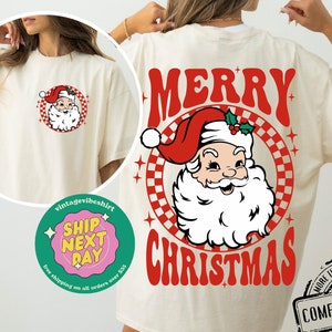 May include: Ivory t-shirt with a red and white checkered circle design featuring a cartoon Santa Claus with a red hat and white beard. The text "Merry Christmas" is printed in red letters below the circle.