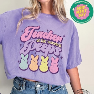 May include: Purple t-shirt with the text "Teacher of the Sweetest Peeps" in a colorful, whimsical font. The shirt features five cartoon Peeps, each a different color: blue, pink, yellow, green, and orange.