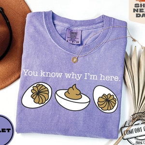 May include: Purple t-shirt with the text "You know why I'm here." and three cartoon images of deviled eggs. The shirt is made of 100% cotton.