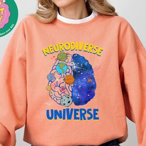 Op de afbeelding: Een terracotta oranje sweatshirt met een afbeelding van een brein met planeten en sterren erin. De tekst "Neurodiverse Universe" is op het sweatshirt gedrukt.