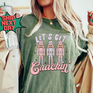 May include: A green t-shirt with the text "Let's Get Crackin'" in pink lettering. Three nutcracker figures are above the text. The shirt has a distressed look and a tag that says "Comfort Colors 100% Cotton".