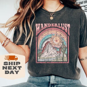 May include: Dark gray Comfort Colors t-shirt with a desert-themed graphic. The design features a rainbow, sun, mountains, and cacti, with the word "WANDERLUST" above. The shirt is 100% cotton and available in more than 10 colors.