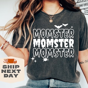 May include: A dark gray t-shirt with the text "Momster" repeated three times in white. The text is in a dripping font style. There is a small spider and a bat graphic above the text.