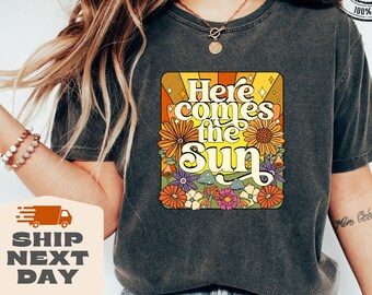 May include: A dark grey t-shirt with a retro-style graphic featuring a sun with flowers and the text "Here comes the sun".
