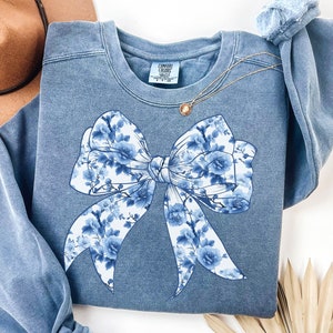 May include: A blue sweatshirt with a white floral bow design. The sweatshirt has a distressed look and is made of a soft, comfortable fabric.