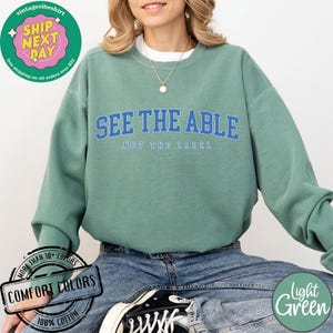 May include: A light green sweatshirt with the text "See the Able, Not the Label" in blue lettering. The sweatshirt is made of 100% cotton and is available in a variety of colours.