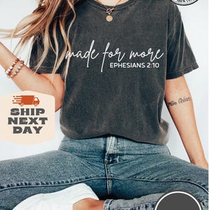 Puede incluir: Una camiseta gris oscuro con el texto "made for more" y "EPHESIANS 2:10" en blanco. La camiseta la lleva puesta una persona con el pelo largo y rubio y unos vaqueros azules.