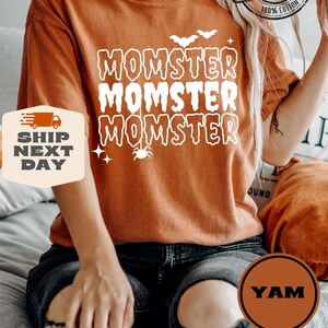 May include: A burnt orange t-shirt with the text "MOMSTER" repeated three times in white. A small spider with a web is hanging below the text. The shirt is on a person wearing black jeans.